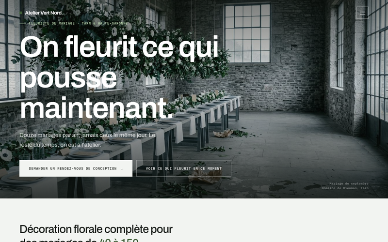 Capture du modèle De saison : une longue table dressée sous une suspension de feuillage et de fleurs blanches, dans un atelier en pierre aux grandes verrières, sous le titre « On fleurit ce qui pousse maintenant. »