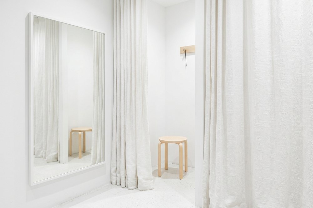 Cabine d’essayage vide : rideau de lin blanc ouvert, tabouret de bois blond, miroir.